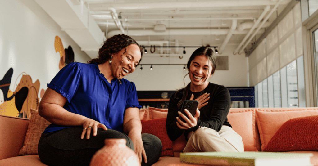 Two women are sitting on a couch looking at a phone and laughing.