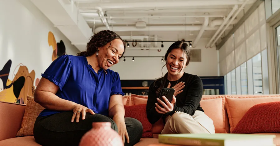 Two women are sitting on a couch looking at a phone and laughing.