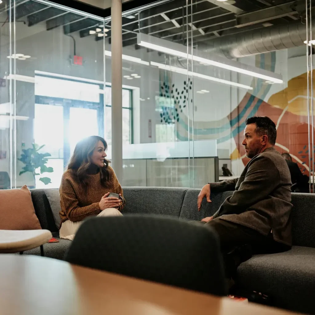 Two individuals are sitting and chatting on a couch in an office.