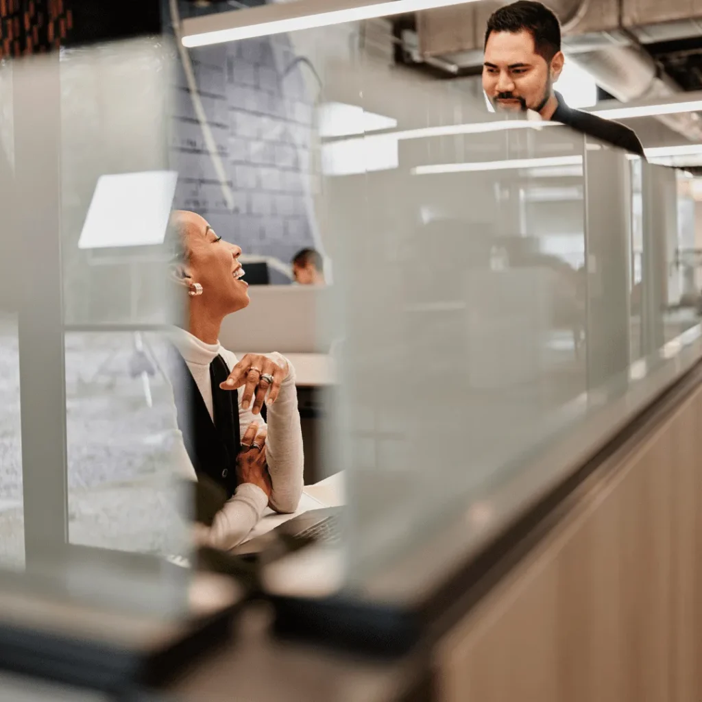 Two coworkers are chatting around a work cubicle.